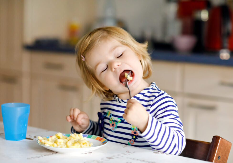 Adorable baby girl eating from spoon noodle, pasta macaroni. Cute healthy toddler child, daughter with spoon sitting in highchair and learning to eat by itself in domestic kitchen or nursery.