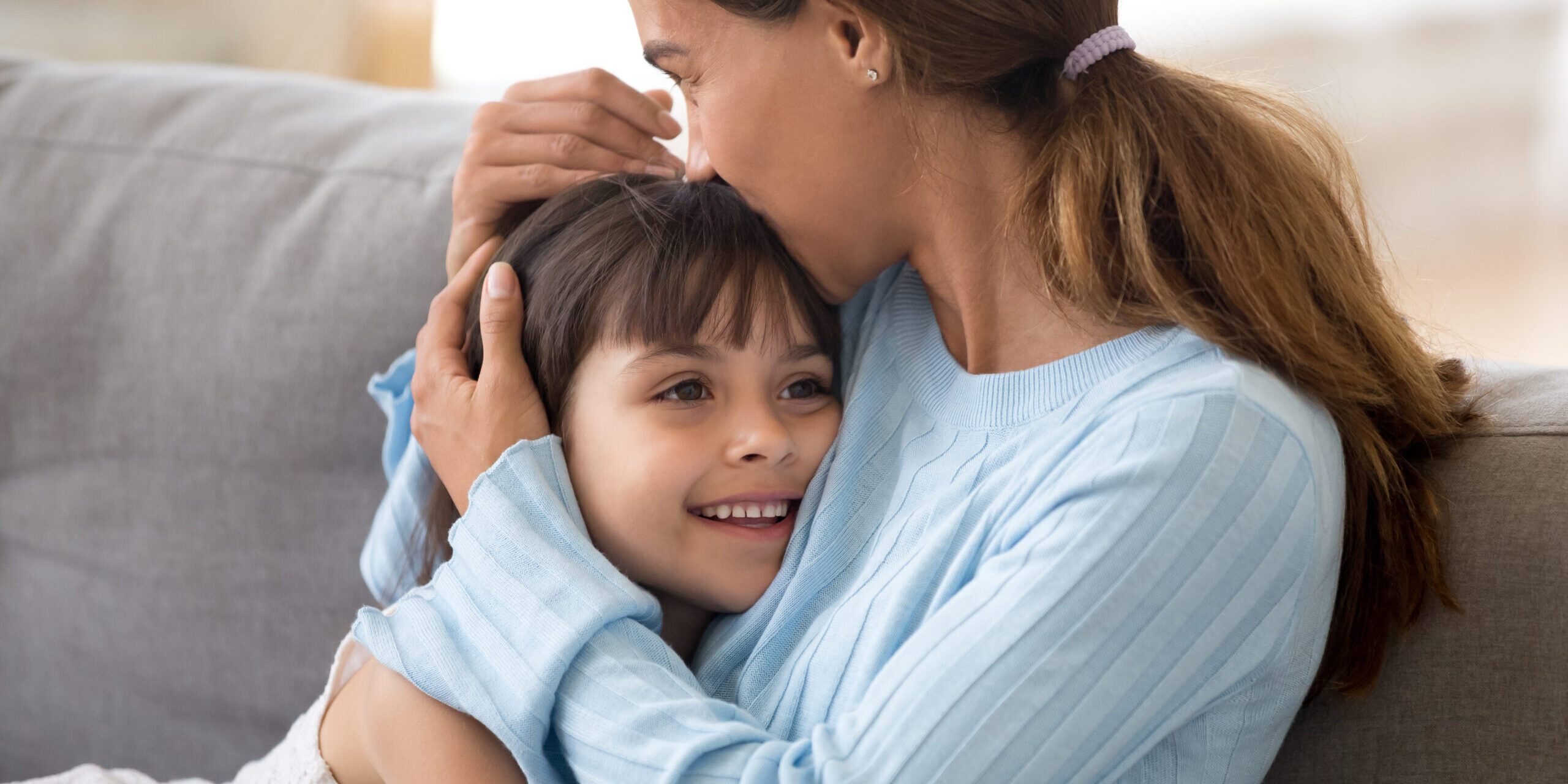 Loving single mother hugging cute little daughter kissing kid sitting on sofa, happy woman mum caressing embracing preschool girl, moms care and support, sincere warm relationships for child concept