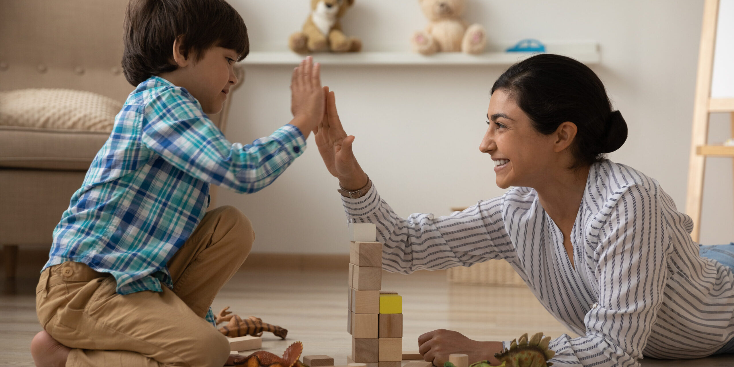 We are great team. Laughing indian mommy give high five to happy kid son celebrate building of high brick tower. Female babysitter construct from brick set with small boy on floor with heating system